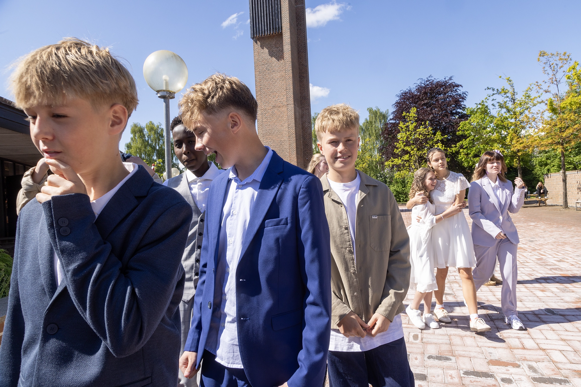 Konfirmander i fint tøj foran et kirketårn på en solskinsdag.